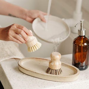 May include: Two wooden dish brushes with natural bristles, one being held and one on a ceramic dish drying rack. A brown glass bottle with a pump dispenser is in the background.