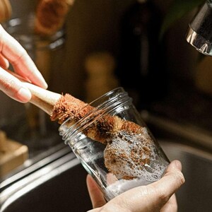 May include: A person is washing a glass jar with a brown and natural bristle bottle brush. The brush is made of natural materials and has a wooden handle. The jar is filled with soapy water and bubbles.