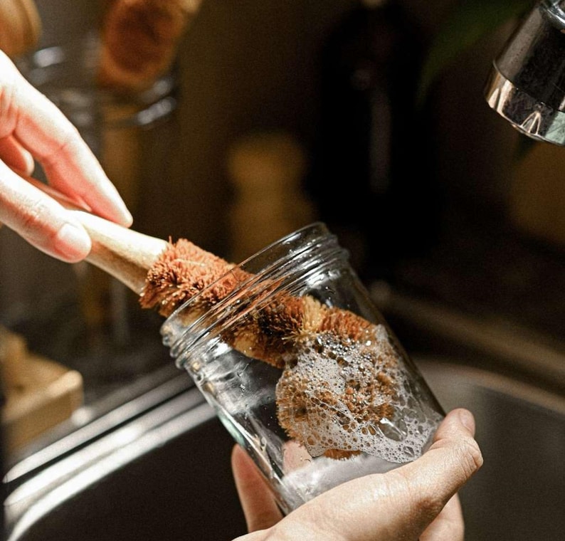 May include: A person is washing a glass jar with a brown and natural bristle bottle brush. The brush is made of natural materials and has a wooden handle. The jar is filled with soapy water and bubbles.