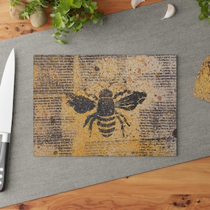 May include: Rectangular cutting board with a distressed, vintage design. The board features a black bee silhouette over a yellow and brown background with faded text. A stainless steel knife is visible on the left.