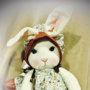 May include: A white plush bunny wearing a brown and white floral dress with a brown bonnet. The bunny has a white face with black eyes and a pink nose.