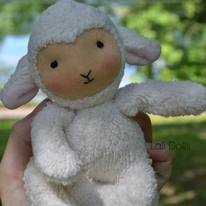 Puede incluir: Un mu&ntilde;eco de peluche de cordero blanco con una textura suave y peluda. El mu&ntilde;eco tiene una expresi&oacute;n dulce e inocente con ojos negros y una peque&ntilde;a nariz rosa. El mu&ntilde;eco est&aacute; siendo sostenido en una mano, y el fondo est&aacute; desenfocado.