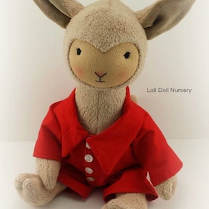 Puede incluir: Un juguete de peluche de cordero con una camisa roja de cuello con botones blancos. El cordero tiene una cara y orejas marrones. El juguete est&aacute; sentado en una superficie blanca. Lali Doll Nursery est&aacute; impreso en el fondo.