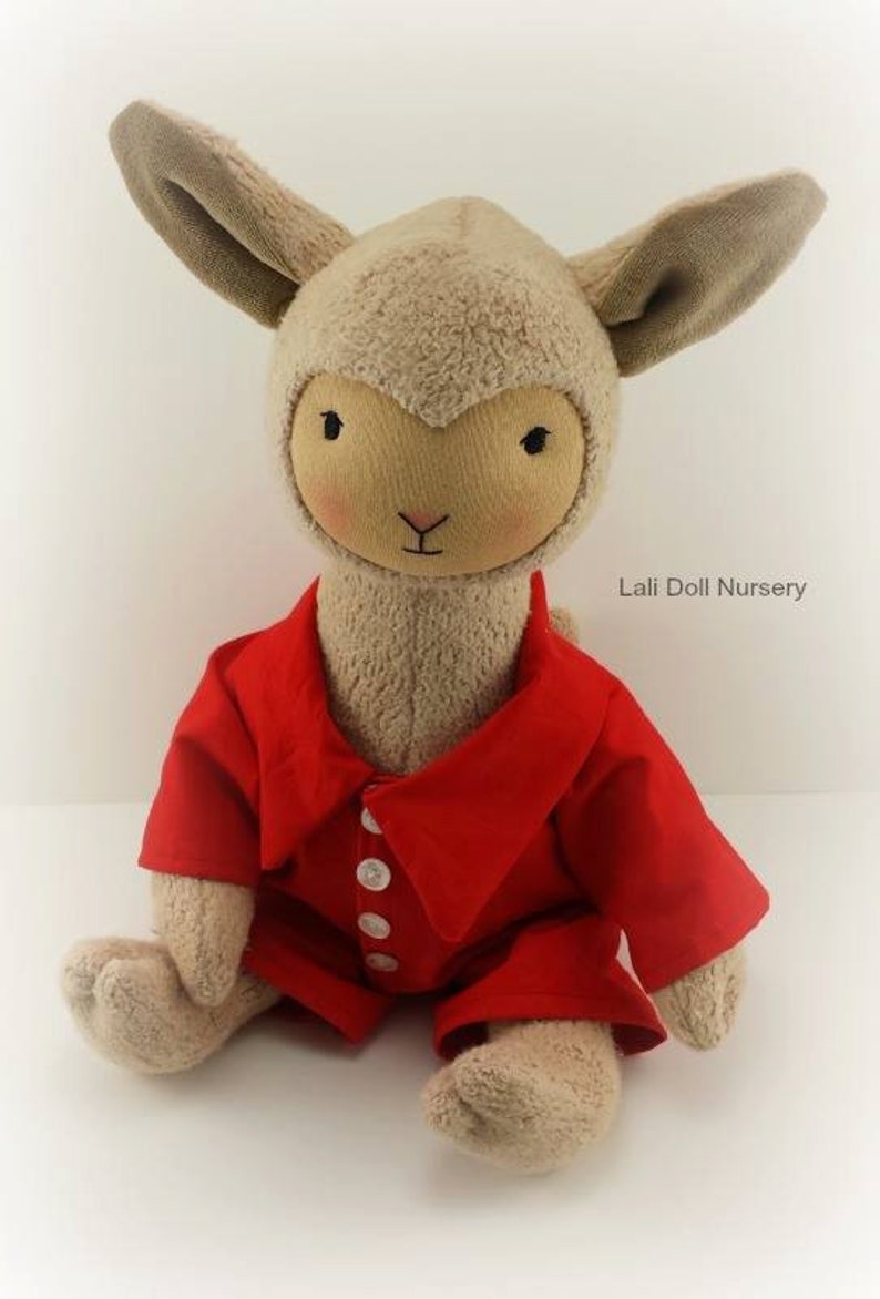 Puede incluir: Un juguete de peluche de cordero con una camisa roja de cuello con botones blancos. El cordero tiene una cara y orejas marrones. El juguete est&aacute; sentado en una superficie blanca. Lali Doll Nursery est&aacute; impreso en el fondo.