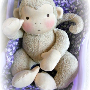 Puede incluir: Un mono de peluche con pelaje marr&oacute;n claro, sentado en una silla con estampado floral morado y blanco. El mono tiene una cara marr&oacute;n claro con mejillas rosadas y un peque&ntilde;o lazo rosa en la cabeza. La mu&ntilde;eca est&aacute; hecha por Lali Doll Nursery.