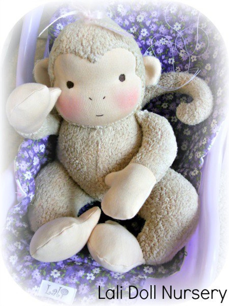 Puede incluir: Un mono de peluche con pelaje marr&oacute;n claro, sentado en una silla con estampado floral morado y blanco. El mono tiene una cara marr&oacute;n claro con mejillas rosadas y un peque&ntilde;o lazo rosa en la cabeza. La mu&ntilde;eca est&aacute; hecha por Lali Doll Nursery.