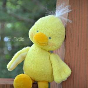 Puede incluir: Un pato de peluche amarillo con ojos azules y un mech&oacute;n de plumas blancas en la cabeza. El pato est&aacute; sentado sobre una superficie de madera.