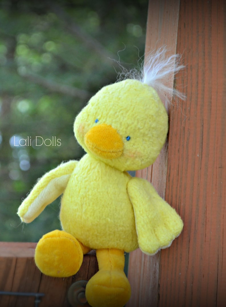Puede incluir: Un pato de peluche amarillo con ojos azules y un mech&oacute;n de plumas blancas en la cabeza. El pato est&aacute; sentado sobre una superficie de madera.