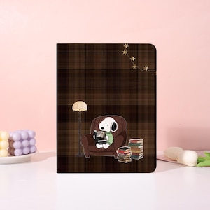 Op de afbeelding: Een bruine geruite notitieboekhoes met een cartoon Snoopy die een boek leest in een bruine fauteuil. Een staande lamp en een stapel boeken zijn ook zichtbaar. De achtergrond is lichtroze.