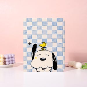 Op de afbeelding: Een rechthoekige, geïllustreerde hoes met Snoopy en Woodstock. De hoes heeft een blauw-witte geblokte achtergrond. Snoopy is wit met zwarte oren en een zwarte neus. Woodstock is geel met een blauwe sjaal. Er is ook een zwarte ster aanwezig.
