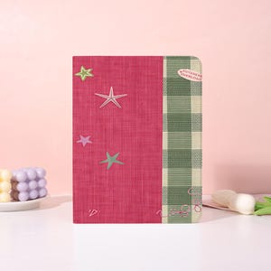 Puede incluir: Un cuaderno rectangular con una cubierta magenta texturizada que presenta varias formas de estrellas en verde claro, rosa y blanco. El lado derecho tiene una banda vertical a cuadros verdes y blancos con una pequeña pegatina ovalada que dice CUTENESS OVERLOAD.