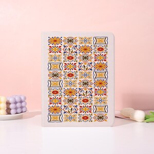 May include: A rectangular notebook with a white cover featuring a repeating vertical pattern of colorful geometric and floral tiles in shades of yellow, red, blue, and dark brown. The design consists of four distinct columns of square motifs.