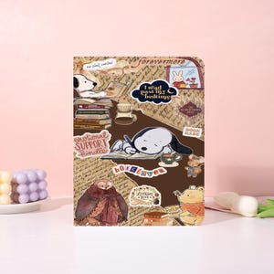 Op de afbeelding: Een bruin notitieboek met diverse cartoonstickers, waaronder Snoopy, Winnie de Poeh en een konijn. Het notitieboek bevat tekst met de tekst "I read past my bedtime", "emotional support kindle" en "literary love".