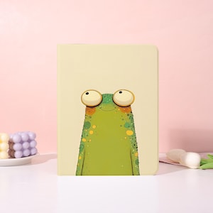 Op de afbeelding: Een rechthoekig notitieboek met een crèmekleurige kaft met een gecentreerde illustratie van een groene kikker met grote, uitpuilende ogen, een kleine glimlach en gele vlekken op het lichaam. De kikker heeft een textuur met fijne lijntjes.