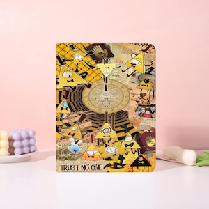 May include: A rectangular notebook featuring a yellow and brown design with cartoon characters and the text "TRUST NO ONE". The cover has a collage of triangles, eyes, and other symbols. The notebook is on a white surface.