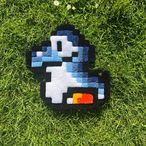 May include: A pixelated rug in the shape of a blue and white dinosaur character with a black outline. The rug has a white belly and a tail with orange and yellow accents. The rug is placed on a bed of green grass with small white flowers.