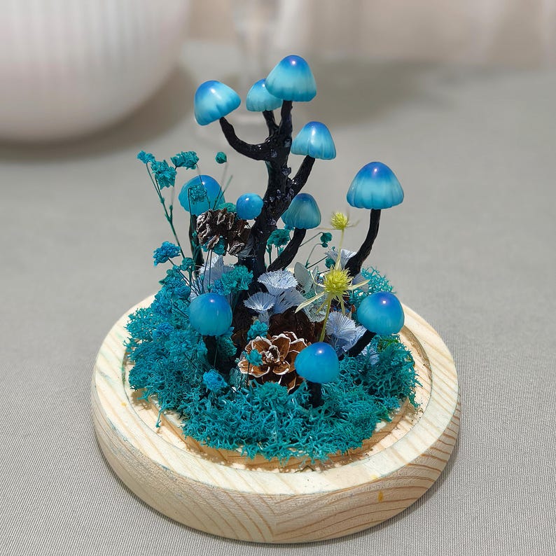 May include: A miniature forest scene in a glass dome with blue mushrooms, green moss, and dried flowers. The base is made of wood.