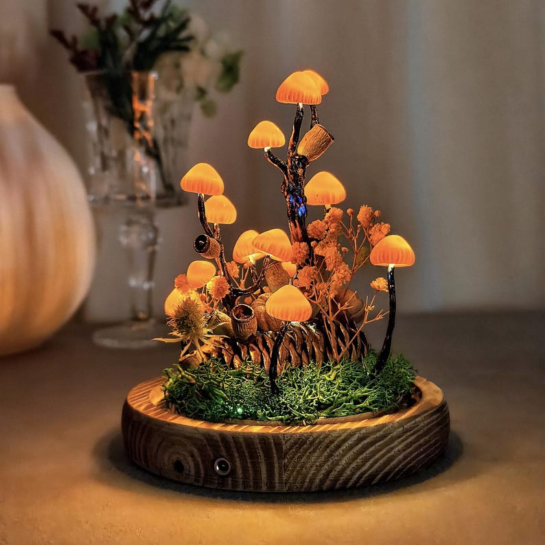 May include: A decorative mushroom lamp with glowing orange caps and a wooden base. The lamp features a miniature forest scene with moss, dried plants, and small details. The warm light creates a cozy ambiance.