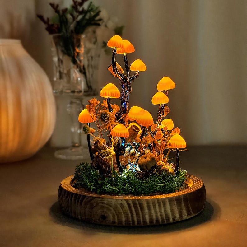 May include: A decorative illuminated mushroom scene. The scene features several glowing orange mushrooms, set amongst dried plants and moss, within a wooden base. The mushrooms are lit with small fairy lights, creating a warm, inviting glow.