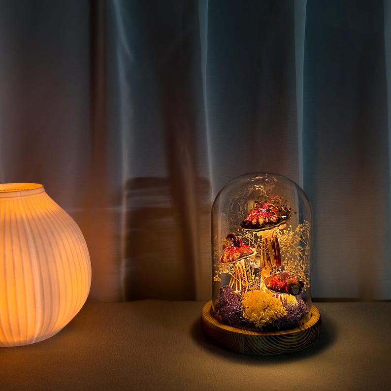 May include: A decorative glass cloche with illuminated red and gold mushrooms on a wooden base. The mushrooms are surrounded by dried flowers and moss. A white, ribbed ceramic lamp is to the left.