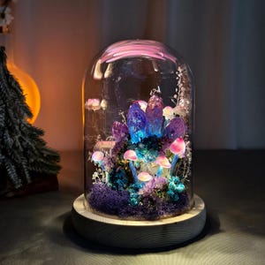 May include: A decorative glass dome featuring a miniature fantasy landscape. Inside, there are glowing pink mushrooms, purple crystals, and blue and purple moss. The dome sits on a wooden base, creating a whimsical and enchanting display.
