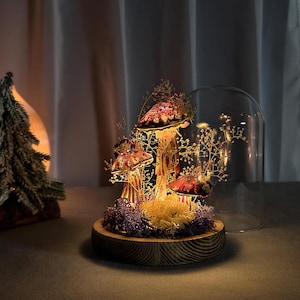 May include: A decorative mushroom display under a glass dome. The mushrooms have red caps with gold stems, surrounded by small lights and dried foliage. The base is a wooden disc, creating a warm, inviting ambiance.