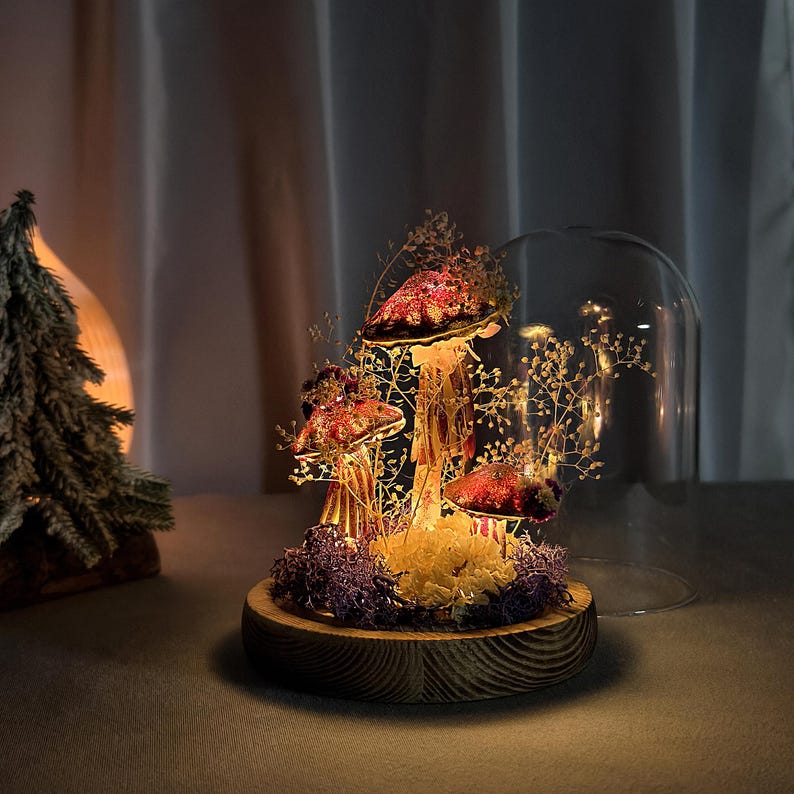 May include: A decorative mushroom display under a glass dome. The mushrooms have red caps with gold stems, surrounded by small lights and dried foliage. The base is a wooden disc, creating a warm, inviting ambiance.