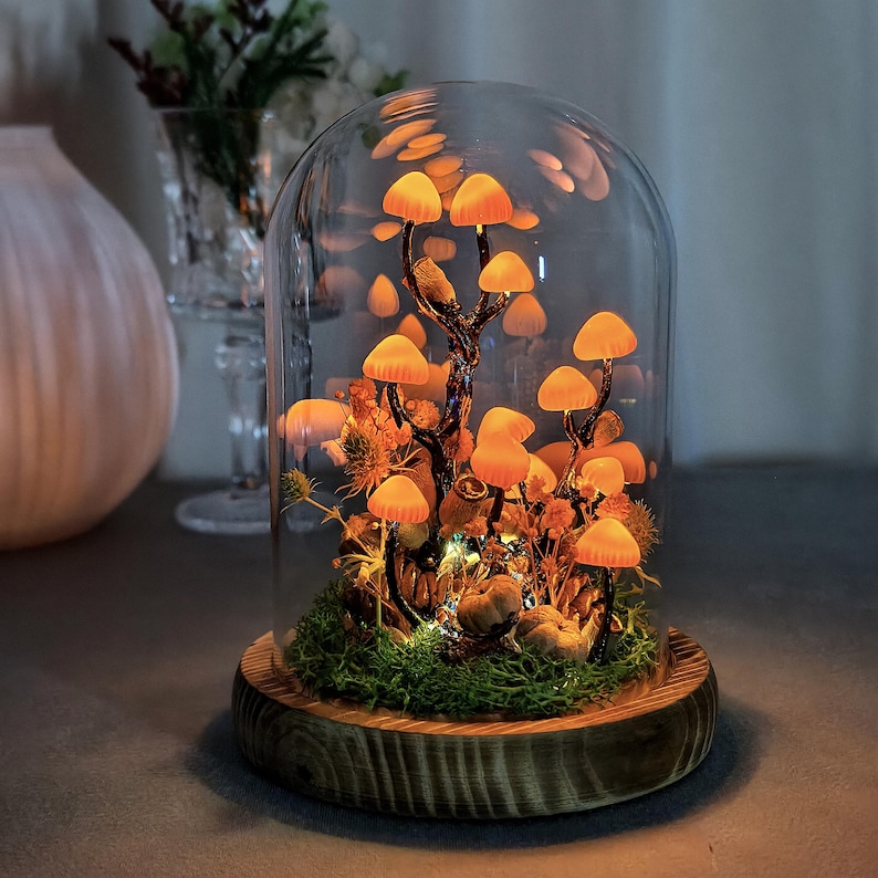 May include: A decorative glass cloche featuring illuminated orange mushroom lights. The mushrooms are arranged on a small tree with dried foliage and moss, set on a wooden base. The cloche is a unique home decor item.