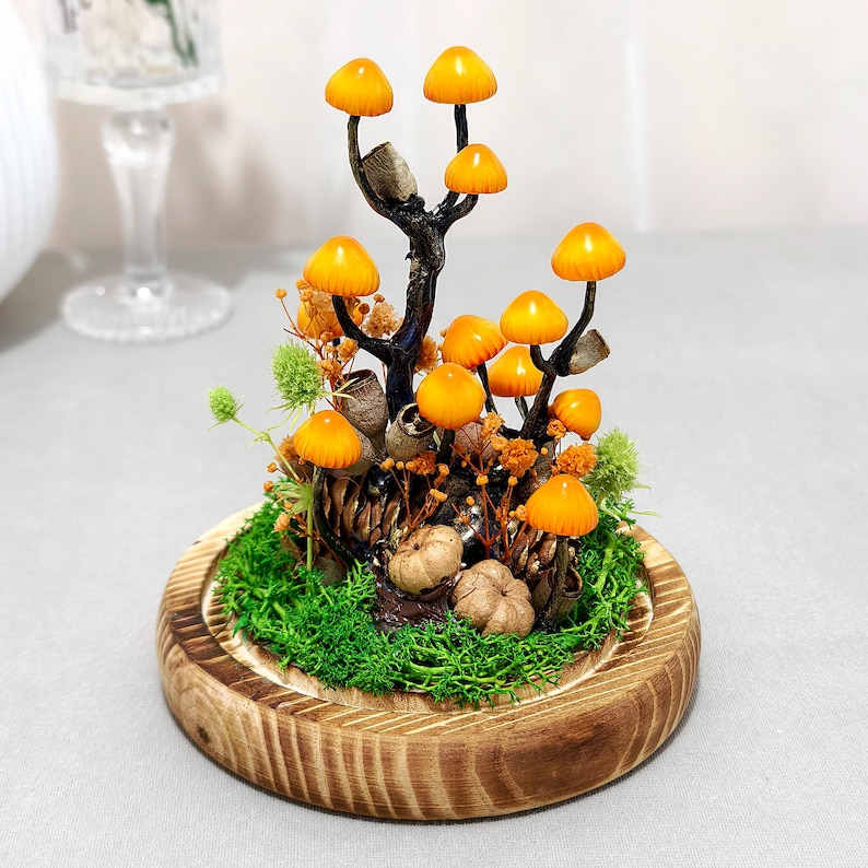May include: A decorative terrarium featuring miniature orange mushroom sculptures with brown stems, set amidst green moss, pinecones, and small pumpkins. The arrangement is housed in a wooden base, creating a whimsical, natural scene.