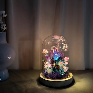 May include: A decorative glass dome with a wooden base houses a miniature fairy garden. The garden features glowing purple crystals, pink mushrooms, and green moss. The dome is lit with a soft, colorful light, creating a whimsical and enchanting display.