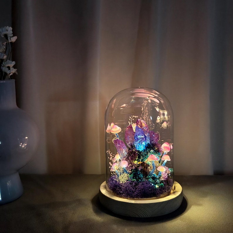 May include: A decorative glass dome with a wooden base houses a miniature fairy garden. The garden features glowing purple crystals, pink mushrooms, and green moss. The dome is lit with a soft, colorful light, creating a whimsical and enchanting display.