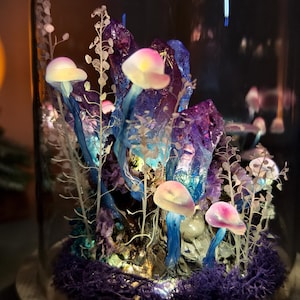 May include: A glass cloche terrarium featuring a miniature fantasy landscape. The scene includes glowing mushrooms with pink and yellow caps, blue and purple crystals, and delicate white foliage. The base is covered in purple moss.