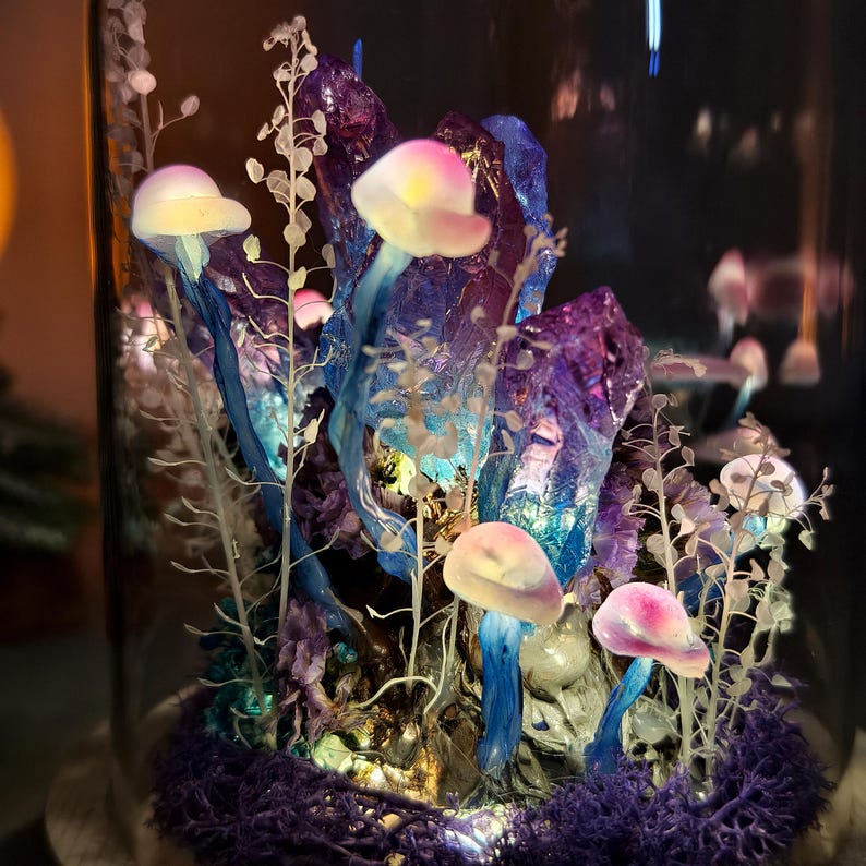 May include: A glass cloche terrarium featuring a miniature fantasy landscape. The scene includes glowing mushrooms with pink and yellow caps, blue and purple crystals, and delicate white foliage. The base is covered in purple moss.
