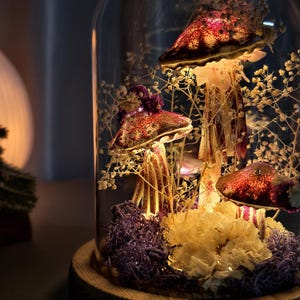 May include: A decorative glass cloche featuring illuminated mushroom sculptures. The mushrooms have burgundy caps with speckled details and are surrounded by dried flowers and foliage. The base is made of wood.