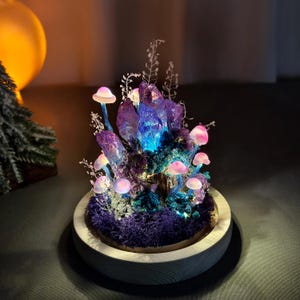 May include: A decorative illuminated mushroom and crystal sculpture. The sculpture features glowing pink mushrooms, purple crystals, and blue and purple moss, all set in a wooden base. The crystals emit a blue light, creating a magical ambiance.