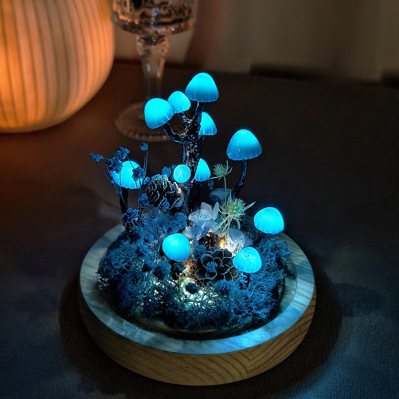 May include: A glowing blue mushroom forest scene in a wooden bowl. The mushrooms are arranged in a cluster with moss and other greenery. The scene is lit from below, creating a magical and whimsical effect.