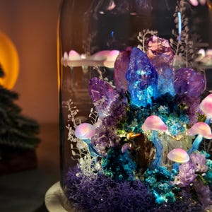 May include: A glass cloche displays a miniature fantasy scene. Purple and blue crystals are illuminated by a blue light. Pink mushrooms and purple moss are also featured. The scene is set on a white base.