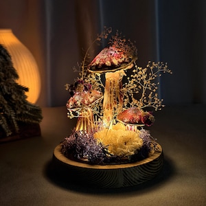 May include: A decorative mushroom-themed lamp with a wooden base. The lamp features several illuminated mushroom-shaped structures with red and brown caps, surrounded by dried flowers and moss. The warm light creates a cozy ambiance.