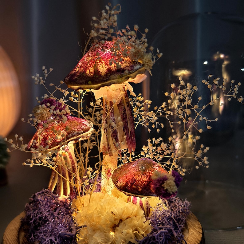 May include: A decorative mushroom arrangement with illuminated caps in shades of red, purple, and gold. The mushrooms are adorned with delicate white floral accents and sit atop a bed of purple and yellow dried flowers. The arrangement is displayed in a glass dome.
