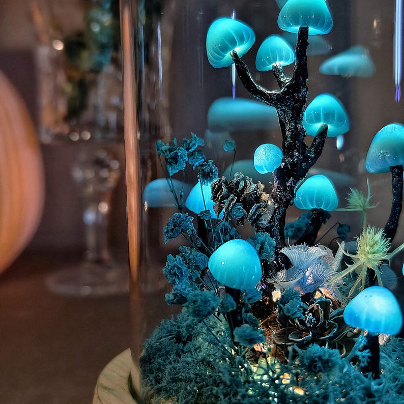 May include: A glass jar terrarium with a glowing blue mushroom forest. The mushrooms are arranged on a black branch with green moss and other plants. The jar is on a wooden base.