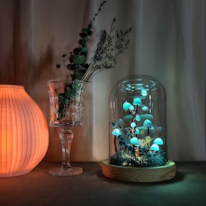 May include: A glass dome with a wooden base containing a glowing blue mushroom forest. The mushrooms are arranged in a cluster and are illuminated from within. The dome is on a table with a vase of dried flowers and a lamp in the background.