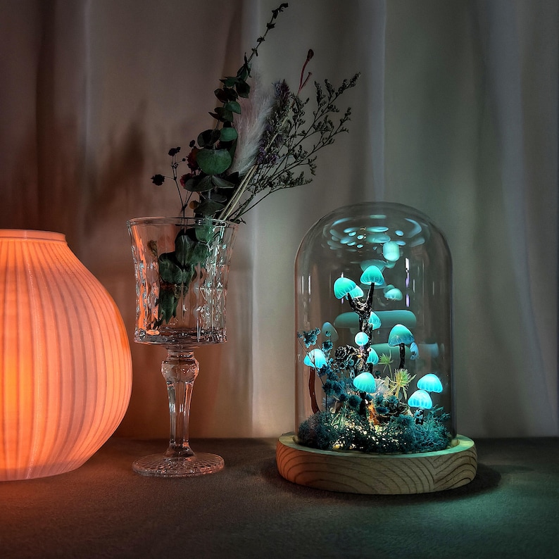 May include: A glass dome with a wooden base containing a glowing blue mushroom forest. The mushrooms are arranged in a cluster and are illuminated from within. The dome is on a table with a vase of dried flowers and a lamp in the background.