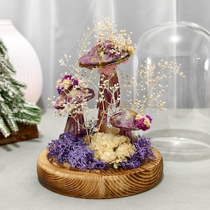 May include: A decorative mushroom arrangement under a glass dome. The mushrooms have purple caps with gold glitter and are surrounded by dried flowers and purple moss. The base is a round, wooden disc.