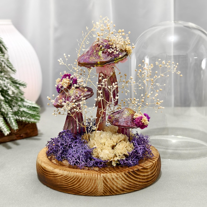 May include: A decorative mushroom arrangement under a glass dome. The mushrooms have purple caps with gold glitter and are surrounded by dried flowers and purple moss. The base is a round, wooden disc.