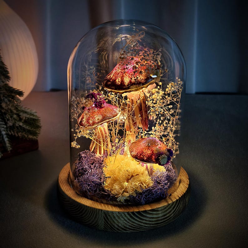 May include: A decorative glass cloche featuring three illuminated, glitter-covered mushroom figurines. The mushrooms are surrounded by dried flowers and moss, set on a wooden base. The cloche creates a warm, ambient glow, perfect for home decor.
