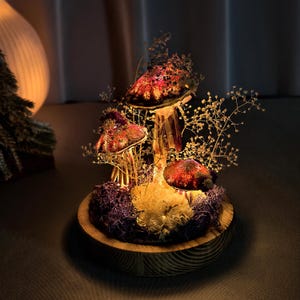 May include: A decorative mushroom light fixture with three illuminated red and brown mushroom caps. The mushrooms are surrounded by dried flowers and moss, set on a wooden base. The light fixture is a unique home decor item.
