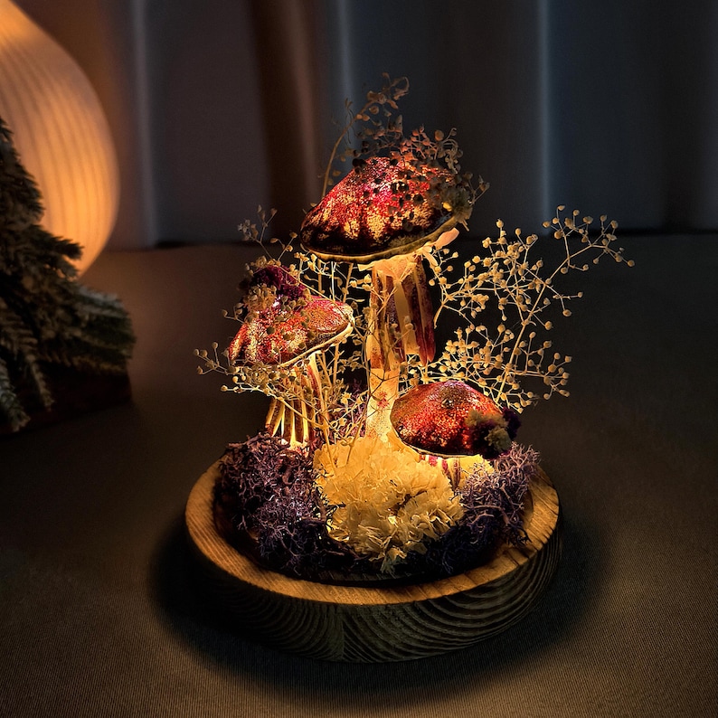 May include: A decorative mushroom light fixture with three illuminated red and brown mushroom caps. The mushrooms are surrounded by dried flowers and moss, set on a wooden base. The light fixture is a unique home decor item.