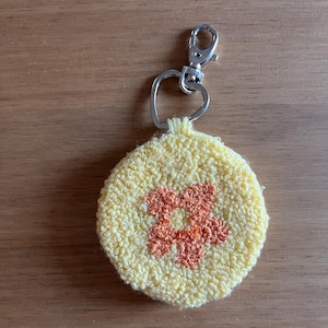 Könnte beinhalten: Ein runder gelber Schlüsselanhänger mit einem kleinen orangefarbenen Blumendesign. Der Schlüsselanhänger hat einen silbernen herzförmigen Ring und einen silbernen Verschluss.