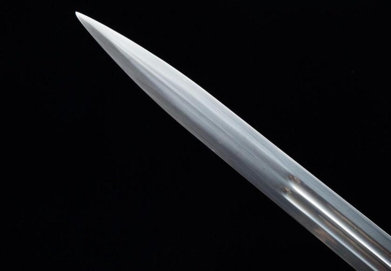 Pu&ograve; includere: Primo piano di una lama di spada argentata e lucidata su sfondo nero. La lama si assottiglia fino a una punta affilata, riflettendo la luce. La superficie della spada presenta sottili scanalature, che suggeriscono un oggetto storico o decorativo.