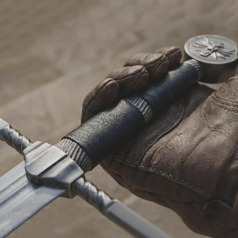 Pu&ograve; includere: Primo piano di una spada con una lama d'argento e un'impugnatura avvolta in pelle nera. L'impugnatura &egrave; tenuta da una mano guantata. &Egrave; visibile anche un medaglione d'argento con un emblema.